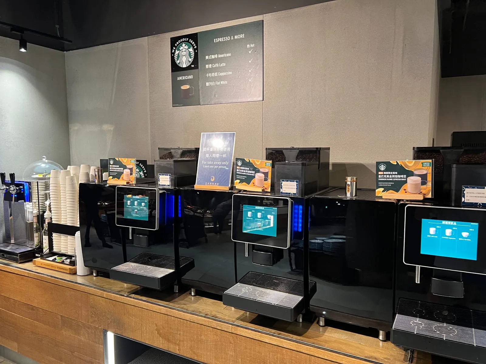 自助咖啡區，三台黑色全自動咖啡機，牆上掛著星巴克 WE PROUDLY SERVE 招牌與熱飲品項看板
