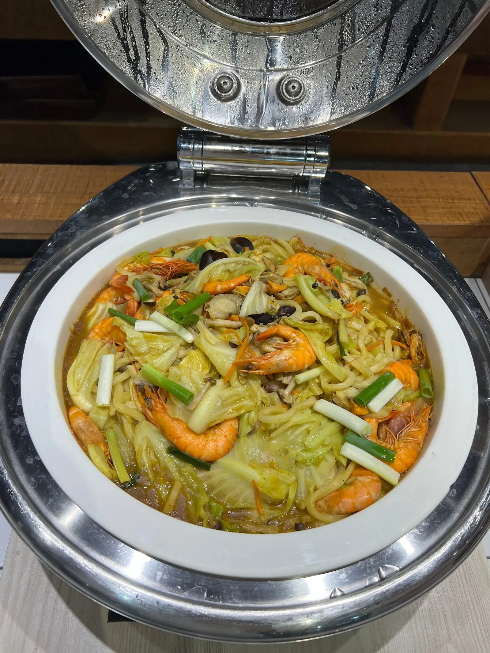 保溫餐檯裡的咖哩海鮮炒烏龍特寫，上面鋪著多隻帶殼鮮蝦、高麗菜與蔥段